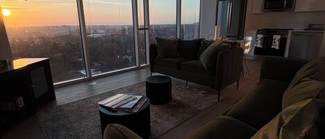 A modern living room with a large window, a sofa, a coffee table, and a kitchen area.