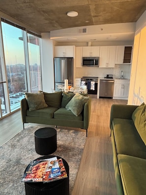 A modern living room with a green sofa, a black coffee table with magazines, and a view of the city through large windows.