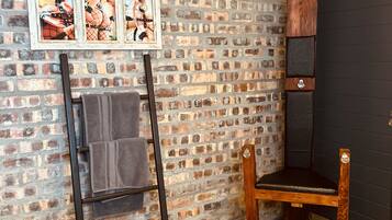 Quarto com parede de tijolos, uma cadeira de madeira, um porta-toalhas de metal e um quadro emoldurado.