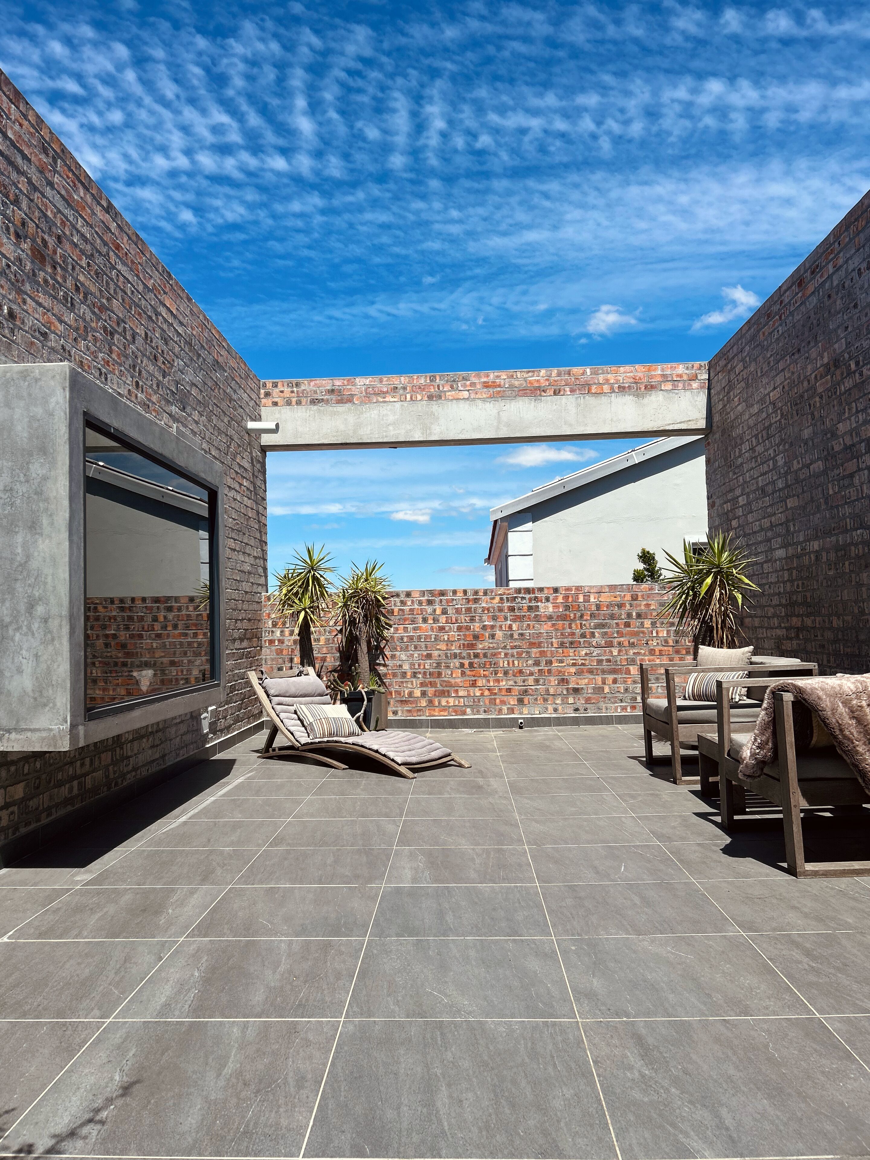 Um pátio externo com parede de tijolos, estrutura de concreto e vista para um edifício através de uma janela.