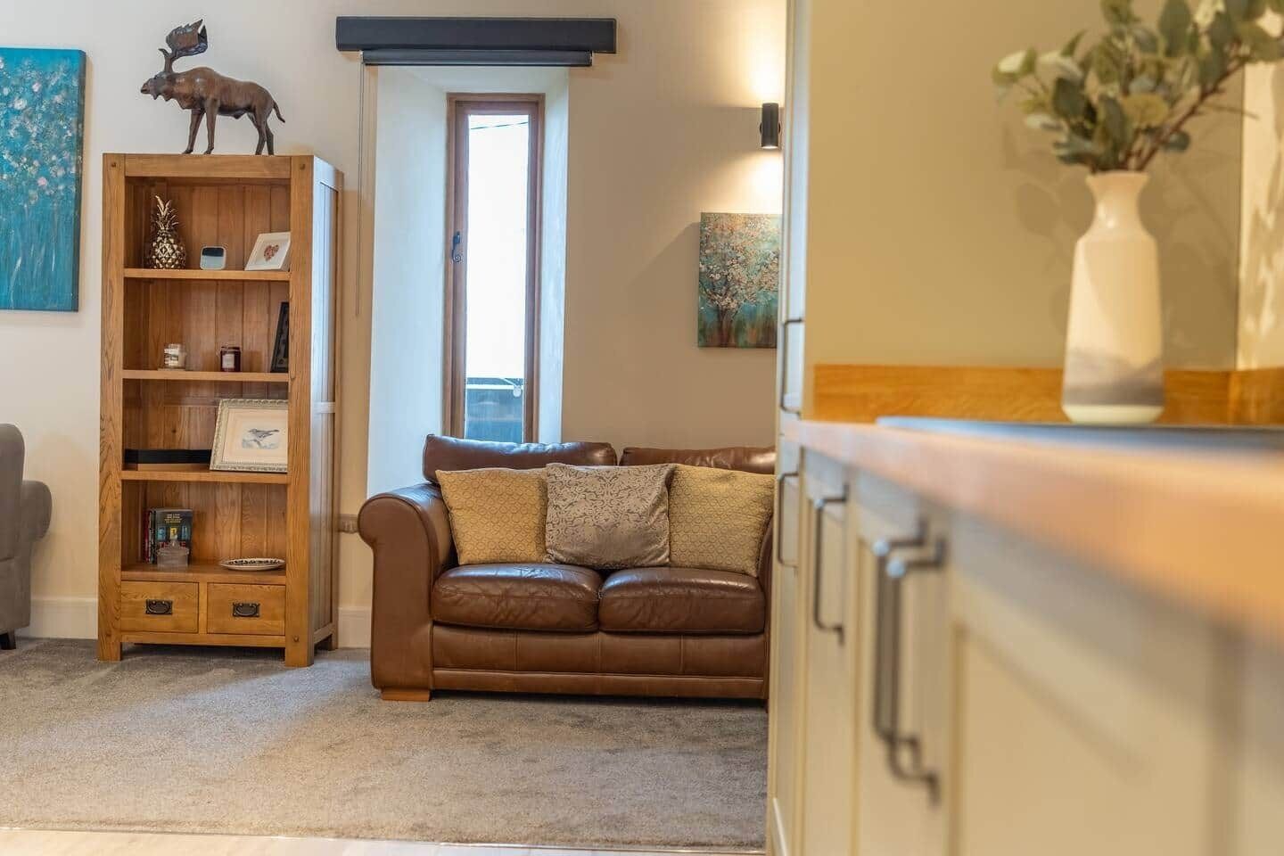 A living room with a brown leather sofa, a wooden bookshelf, and a vase with flowers on a cabinet.