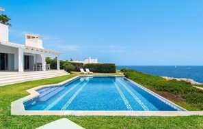 Une piscine avec vue sur la mer, un bâtiment blanc doté d’un patio couvert et un ciel d’un bleu limpide.