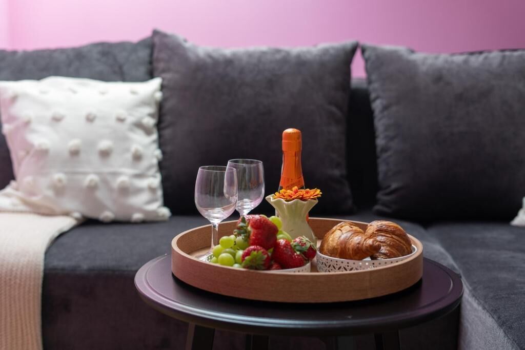 Una bandeja con frutas, una botella y un croissant sobre una mesita frente a un sofá con cojines oscuros.