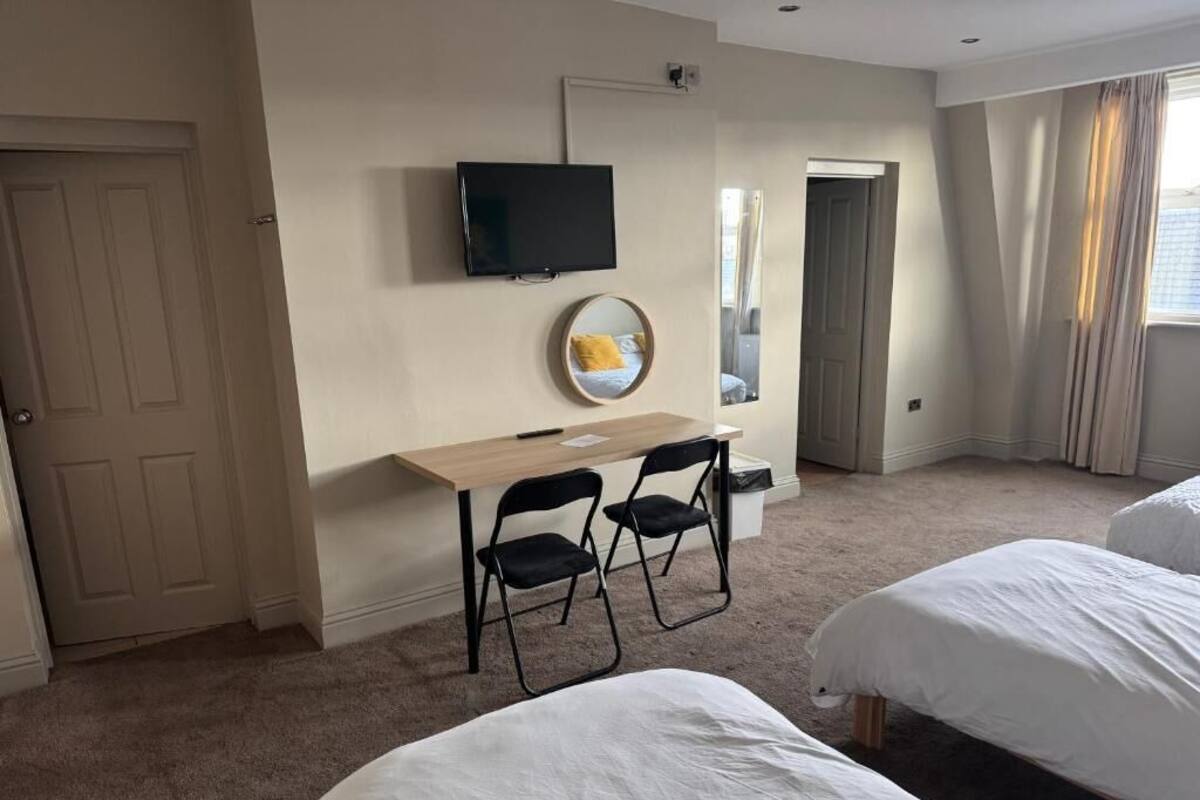 Quarto de hotel com duas camas, uma escrivaninha com cadeiras, televisão e um espelho.