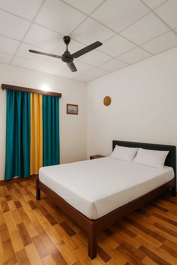 A hotel room with a bed, wooden flooring, a ceiling fan, a window with curtains, and a wall clock.
