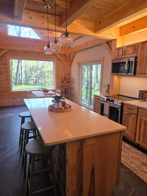 A spacious kitchen with wooden cabinets, a large island with stools, and a view of the outdoors through large windows. - Tranquil cabin in Somerset PA (Somerset)