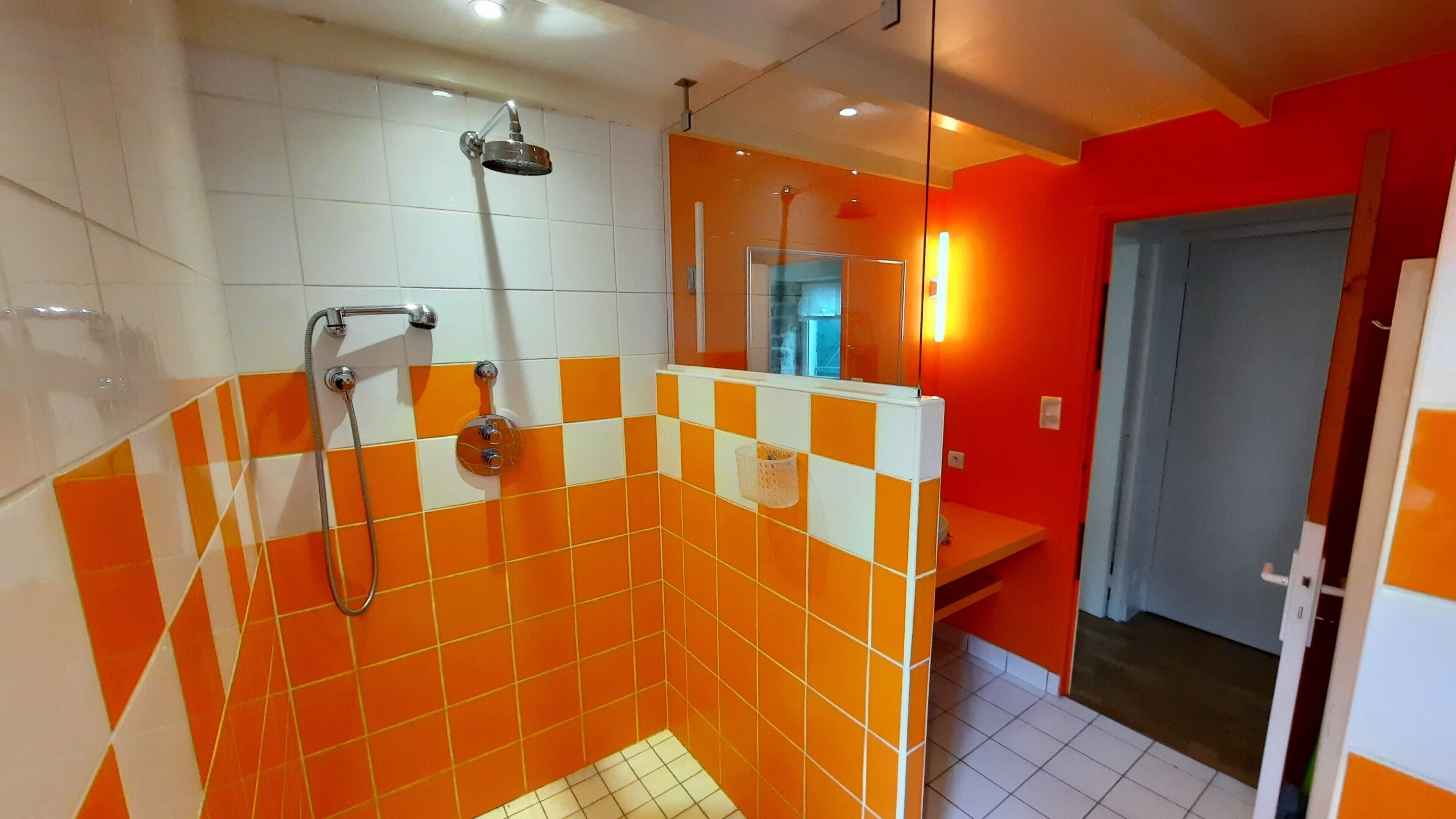 Une salle de bain avec des carreaux orange et blancs, une douche équipée d’un pommeau de douche amovible et un miroir.