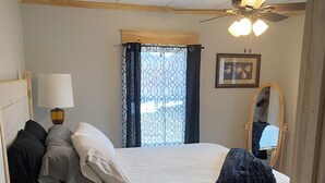 Een slaapkamer met een bed, nachtkastje, lamp, raam met gordijnen, plafondventilator en een spiegel.
