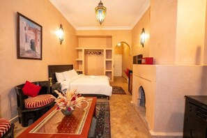 A hotel room with a bed, a sofa, a table with a vase, a fireplace, and a hallway leading to another room. - Mumtaz Dar - Suite Deluxe 3 (Ouahat Sidi Brahim)