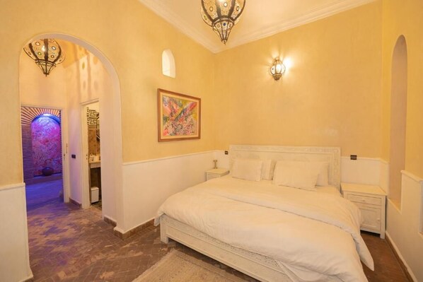 A bedroom with a large bed, two bedside tables, a nightstand, a painting on the wall, and an arched doorway leading to another room.