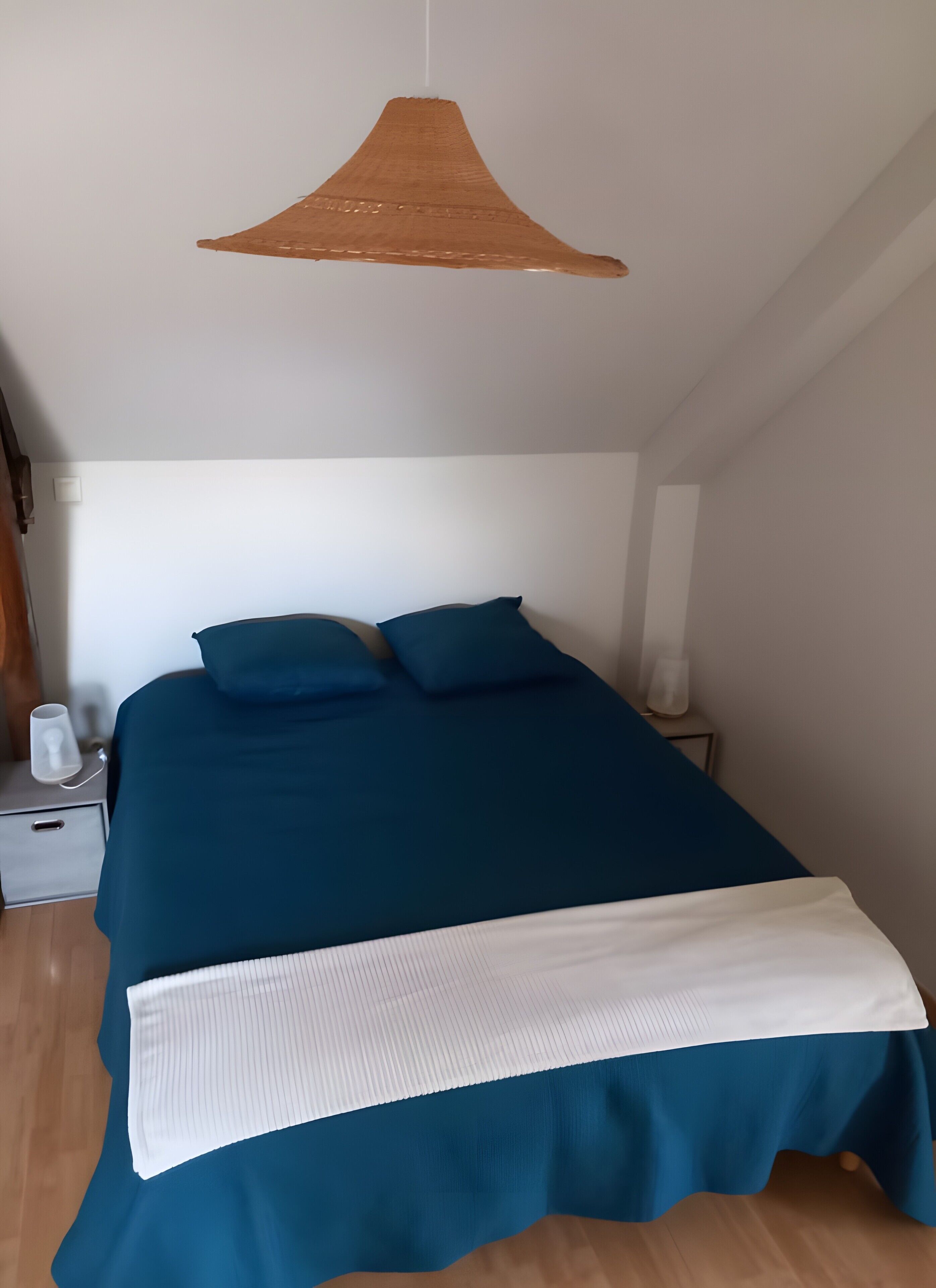 Un lit avec une couvre-lit bleue et des oreillers blancs, un plaid blanc, une table de chevet avec une lampe et un luminaire suspendu.