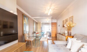 A modern living room with a glass dining table, comfortable seating, a flat-screen TV, and a sofa with decorative pillows.