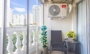 A balcony with a white balustrade, a grey lounge chair, a potted plant, a small table with a vase of sunflowers, and an air conditioning unit. - 3-bedroom Apt in Setor Bueno Pdaa0304 (Goiânia)