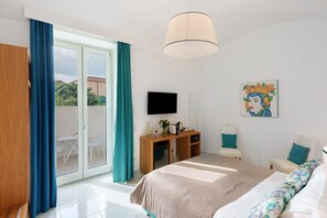 A hotel room with a bed, a desk, a chair, a TV, and a sliding glass door to the outside. - Sorrento Stylish – Deluxe Room with Balcony (Sorrento)