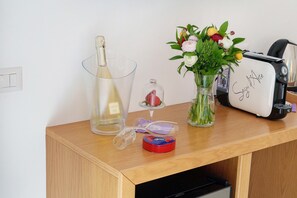 A wooden table with a bottle, a glass, a vase with flowers, a toaster, and a small red tin. - Sorrento Stylish – Deluxe Room with Balcony (Sorrento)