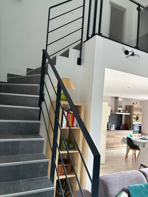 A staircase with black railings leading to a second floor, a bookshelf with books, a dining area with a table and chairs, and a kitchen area with cabinets and appliances. - Villa 'Corinne' with Private Terrace, Wi-Fi and Air Conditioning (Sainte-Marie-la-Mer)