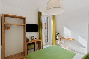 Une chambre d’hôtel comprenant un lit, un bureau avec une chaise, une télévision, une fenêtre avec des rideaux et une porte avec un porte-manteau.