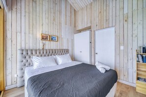A bedroom with a tufted headboard, a large bed with a grey quilt, two white doors, and a wooden bookshelf. - Bluelight Concón- Sea View w/ Wifi (Concón)