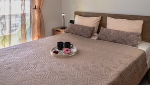 Una cama bien hecha con una bandeja que tiene dos tazas y una dona, una mesita de noche con una lámpara y una ventana con cortinas.