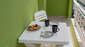 Un pequeño espacio al aire libre con mesa y sillas blancas, un plato de pasteles y tazas negras.