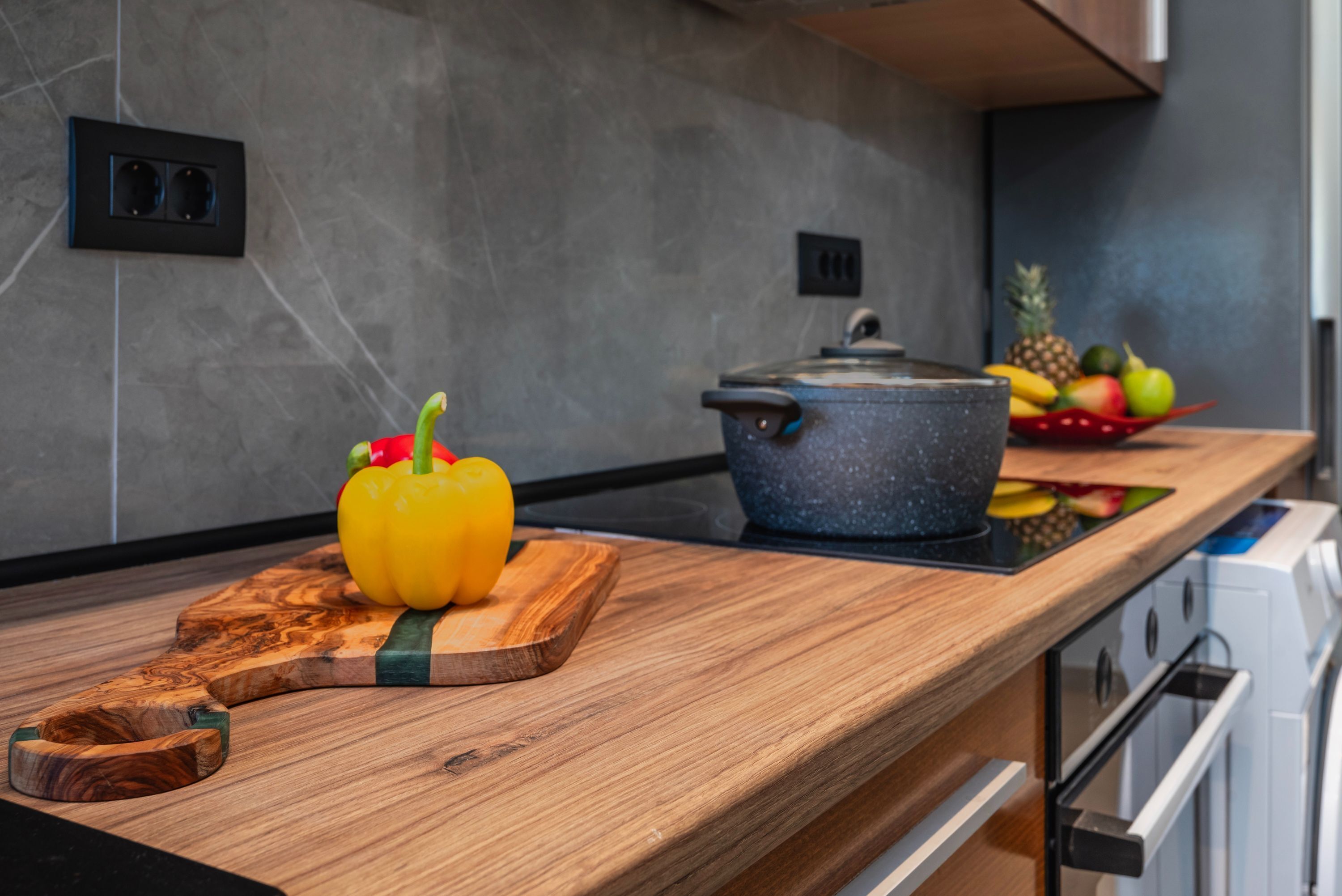 A kitchen counter with a wooden cutting board, a yellow bell pepper, a pot, and a fruit bowl.
