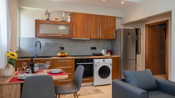 Una cocina moderna con armarios de madera, horno empotrado y lavadora. Hay un comedor con una mesa para dos, un sofá y un florero con flores.
