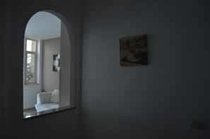 A room with a white armchair, a small table, a framed picture on the wall, and a window with a view of another room. - IL SOGNO MODERNO (Caltagirone)