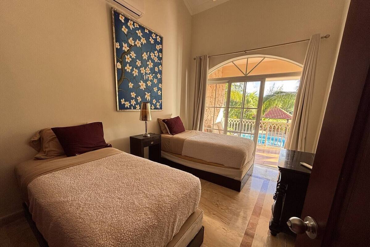 A hotel room with two beds, a nightstand, a lamp, and a large window with a view of a pool and greenery.
