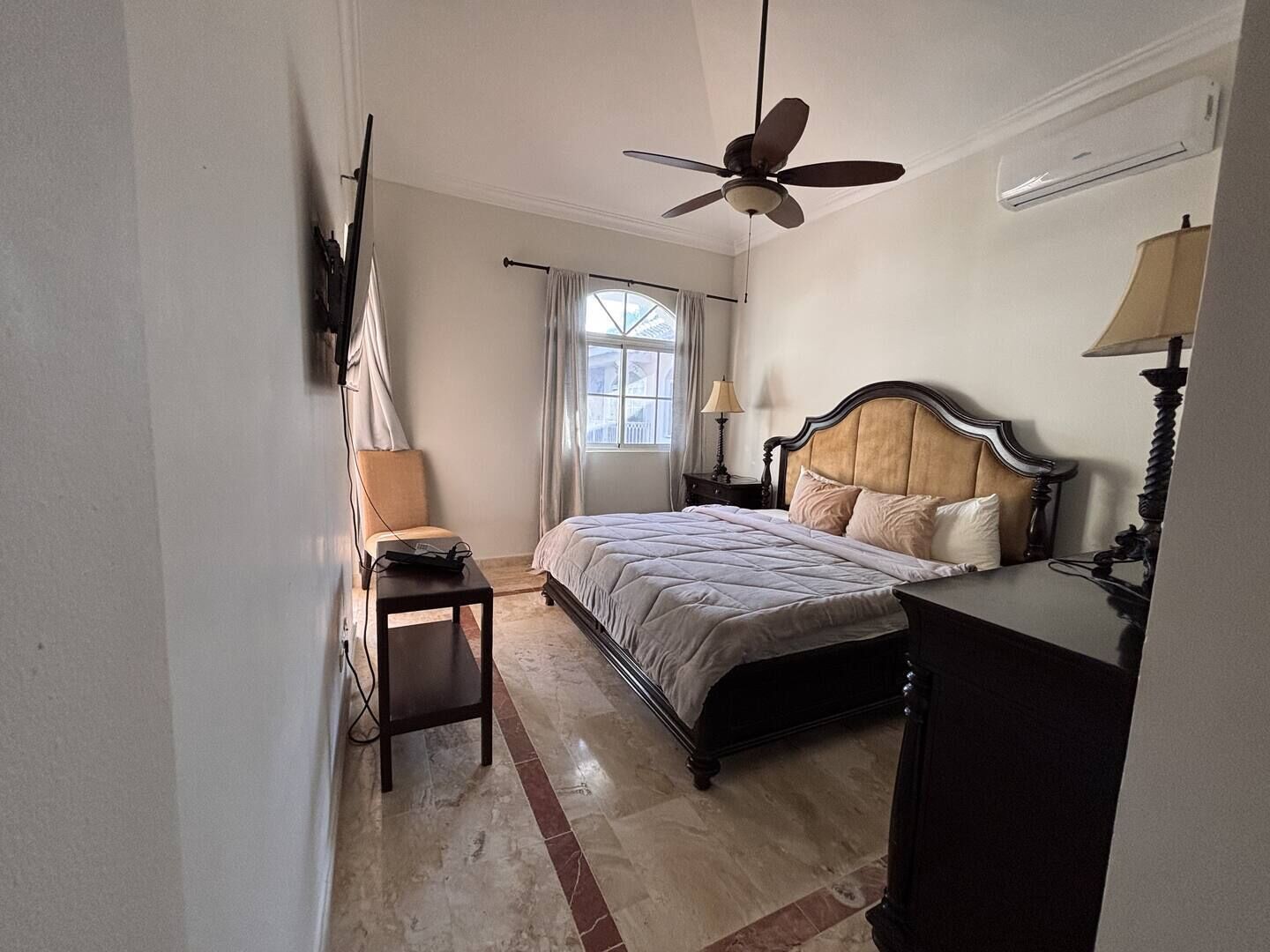 Un dormitorio con cama, escritorio, silla, ventilador de techo y ventana con cortinas.