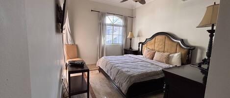 Un dormitorio con cama, escritorio, silla, ventilador de techo y ventana con cortinas.