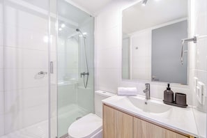 Un baño moderno con ducha envuelta en vidrio, inodoro blanco y tocador con espejo y lavamanos.