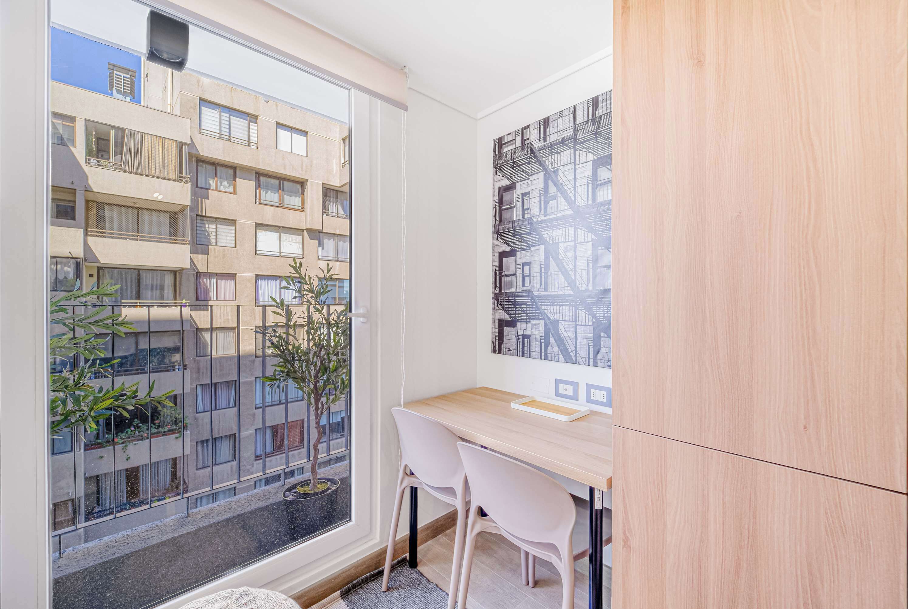 Une petite pièce avec une fenêtre donnant sur un immeuble, un bureau en bois, deux chaises et une plante en pot.
