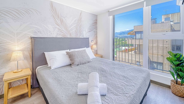 A neatly made bed with a grey headboard, a bedside lamp, and a view of the city through a large window. - Espacio Coventry II - 1D Wifi y AC (Ñuñoa)