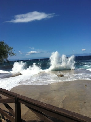 A beach with waves crashing onto the shore, a clear sky, and a wooden railing in the foreground.