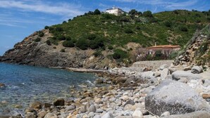 A rocky beach with a white building on a hill, clear blue water, and a small structure with a red roof. - Villa tre fari all'Isola d'Elba (Colle D'orano)