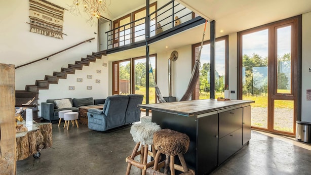 Un salón comedor amplio con techo alto, una escalera que sube a un nivel superior, una isla grande con taburetes y vistas al exterior a través de amplias ventanas.
