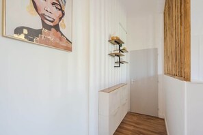 A narrow hallway with a wooden floor, white walls, and a painting on the left. There are shelves with items on the right wall. - Bright, modern 2-bedroom in city center (Hyères)