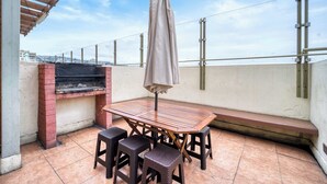 Een terras met een houten tafel, vier krukken en een overdekte parasol.