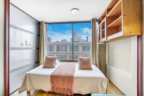 A bedroom with a large window, a bed with a beige blanket, two pillows, and wooden shelving above the bed.