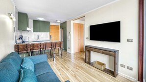 Een moderne woonkamer met een blauwe bank, een houten eettafel met barstoelen, een flatscreen televisie aan de muur en een keuken op de achtergrond.
