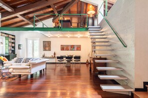 A spacious living area with a wooden floor, a white sofa, a dining table with chairs, and a staircase with a green railing.