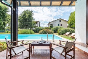 A covered patio with a pool, wooden furniture, and a view of a house and greenery.