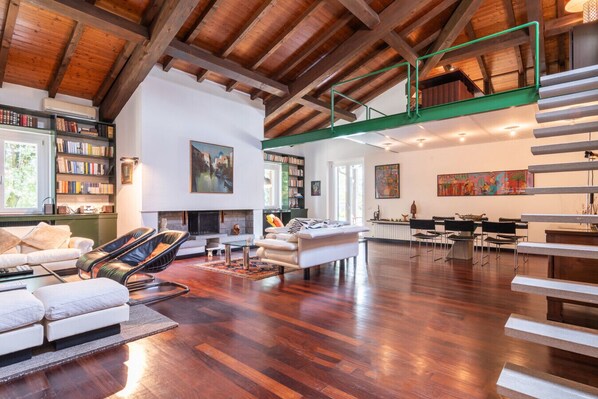 A spacious living area with a high ceiling, wooden beams, a fireplace, and a dining area with chairs and a table.