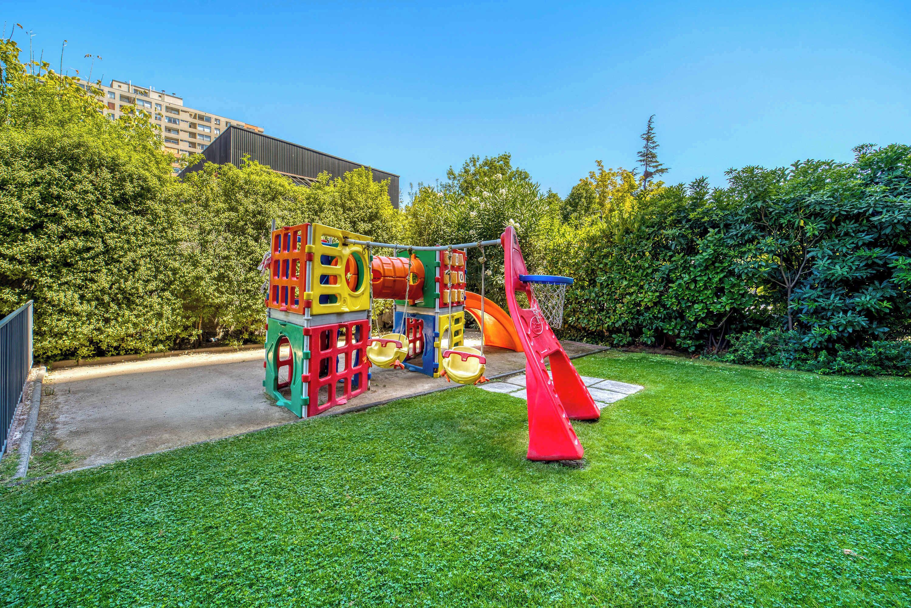 A playground with colorful play structures, including a slide and swings, set in a grassy area with trees and a building in the background.