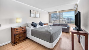 A modern hotel room with a large bed, a wooden nightstand, a TV on a stand, and a city view through the window. - Las Condes Urban Apartment W/ Wifi and Pool (Las Condes)