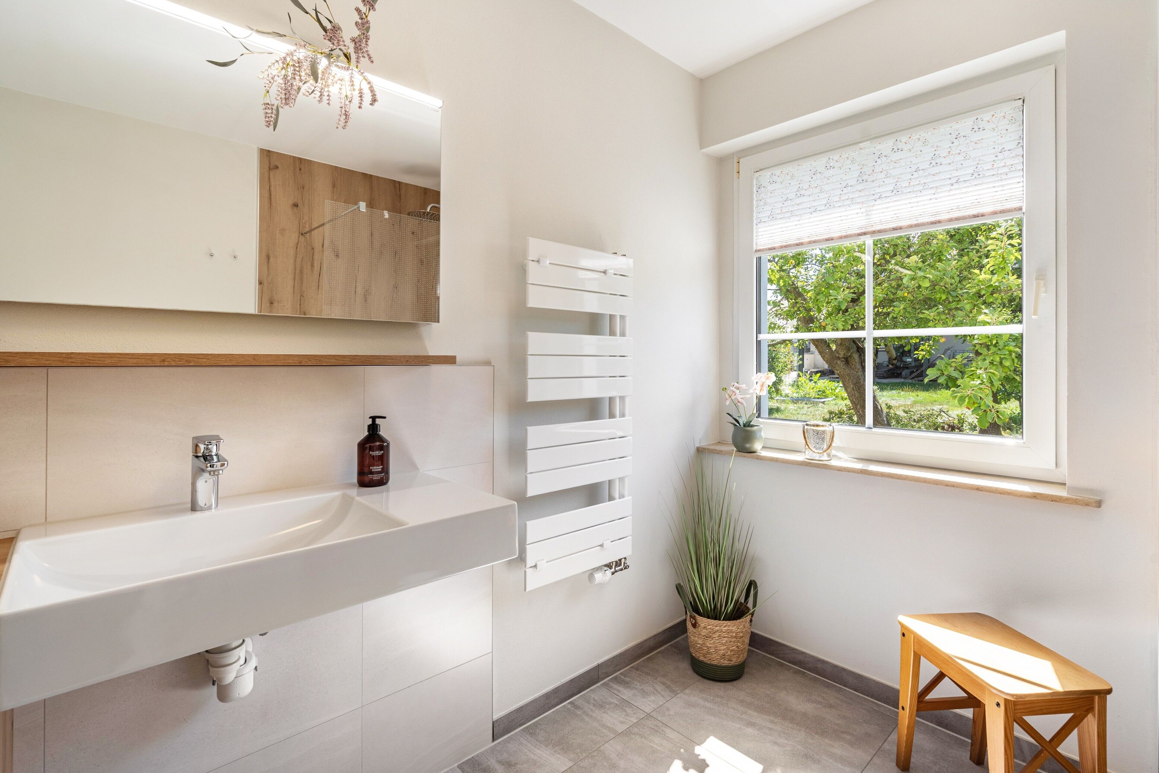A modern bathroom with a large sink, a mirror, a window with a view of greenery, and a wooden stool.
