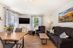 Ein modernes Wohnzimmer mit einem Holzesstisch, einer Couch, einem Sessel und einem an der Wand befestigten Flachbildfernseher.