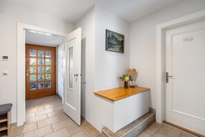 Een gang met een houten deur, een witte muur met een lijstfoto en een klein houten plankje met een vaas.