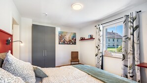Ein Schlafzimmer mit Bett, Kleiderschrank, Stuhl, einem Gemälde und einem Fenster mit Vorhängen.