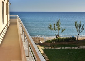 A balcony with a view of the sea, a small garden with trees, and a sandy beach.